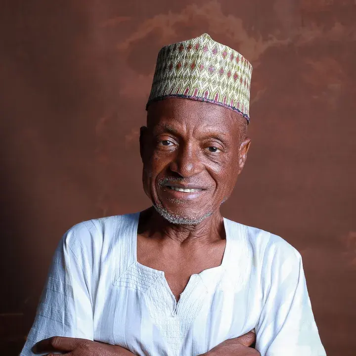 An older man in a white smock and embroidered cap, smiling.