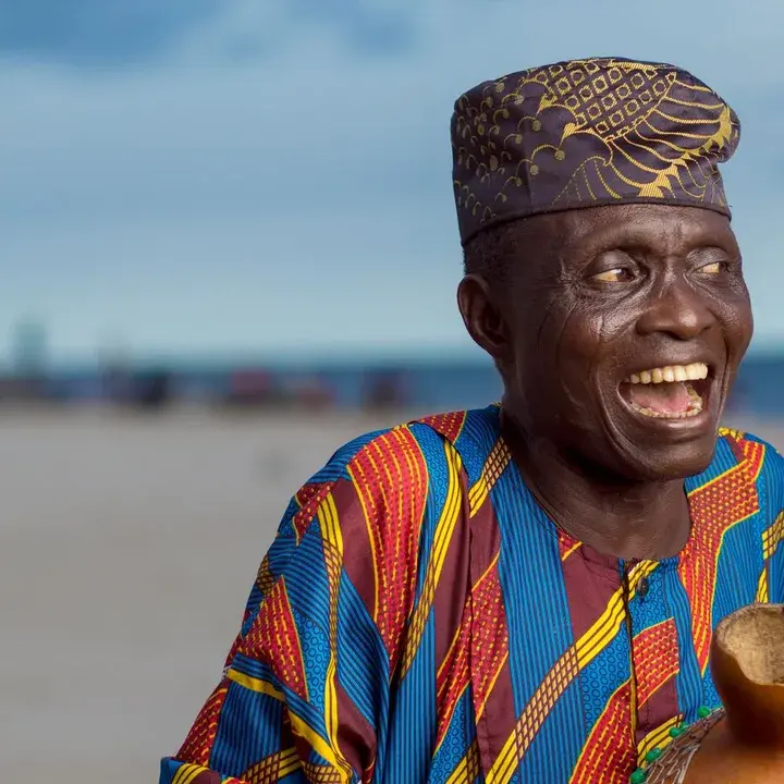 An older man in bright kente cloth, laughing.