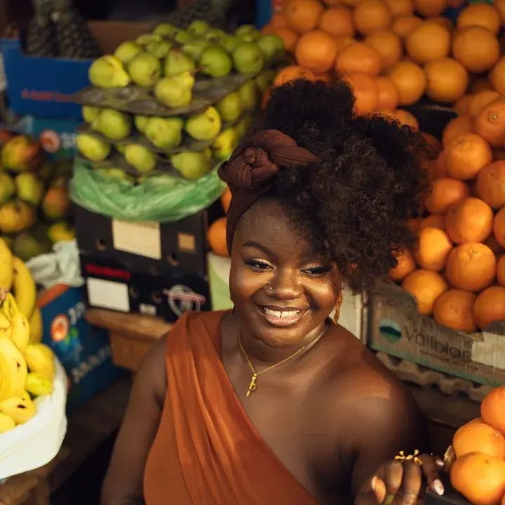 A trader smiling at her fruit stall, oranges and pears stacked behind her.