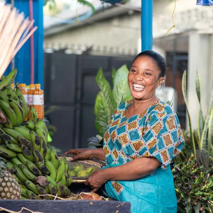 A trader laughing behind a stall of plantain and greens.