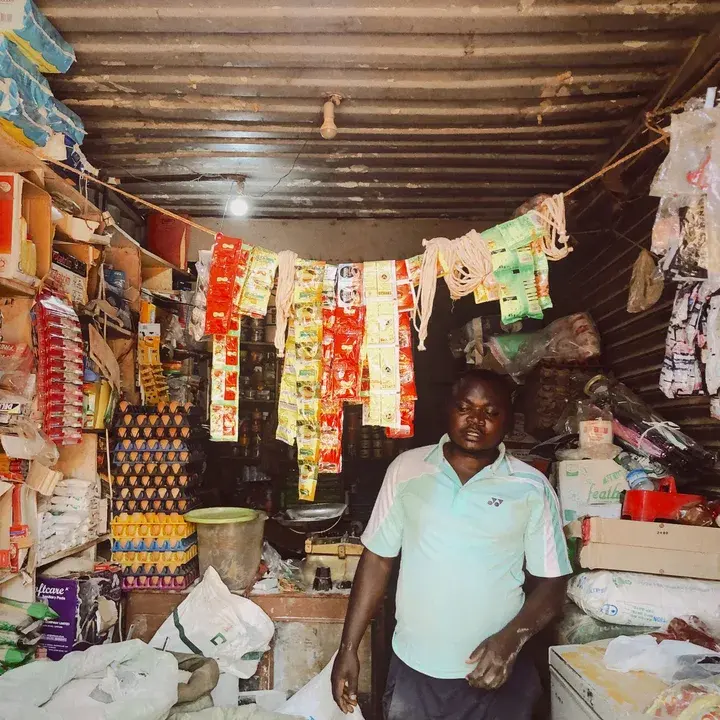 A shopkeeper standing in his provisions shop, goods hung from the ceiling.