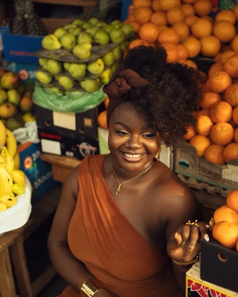 A trader smiling at her fruit stall, oranges and pears stacked behind her.